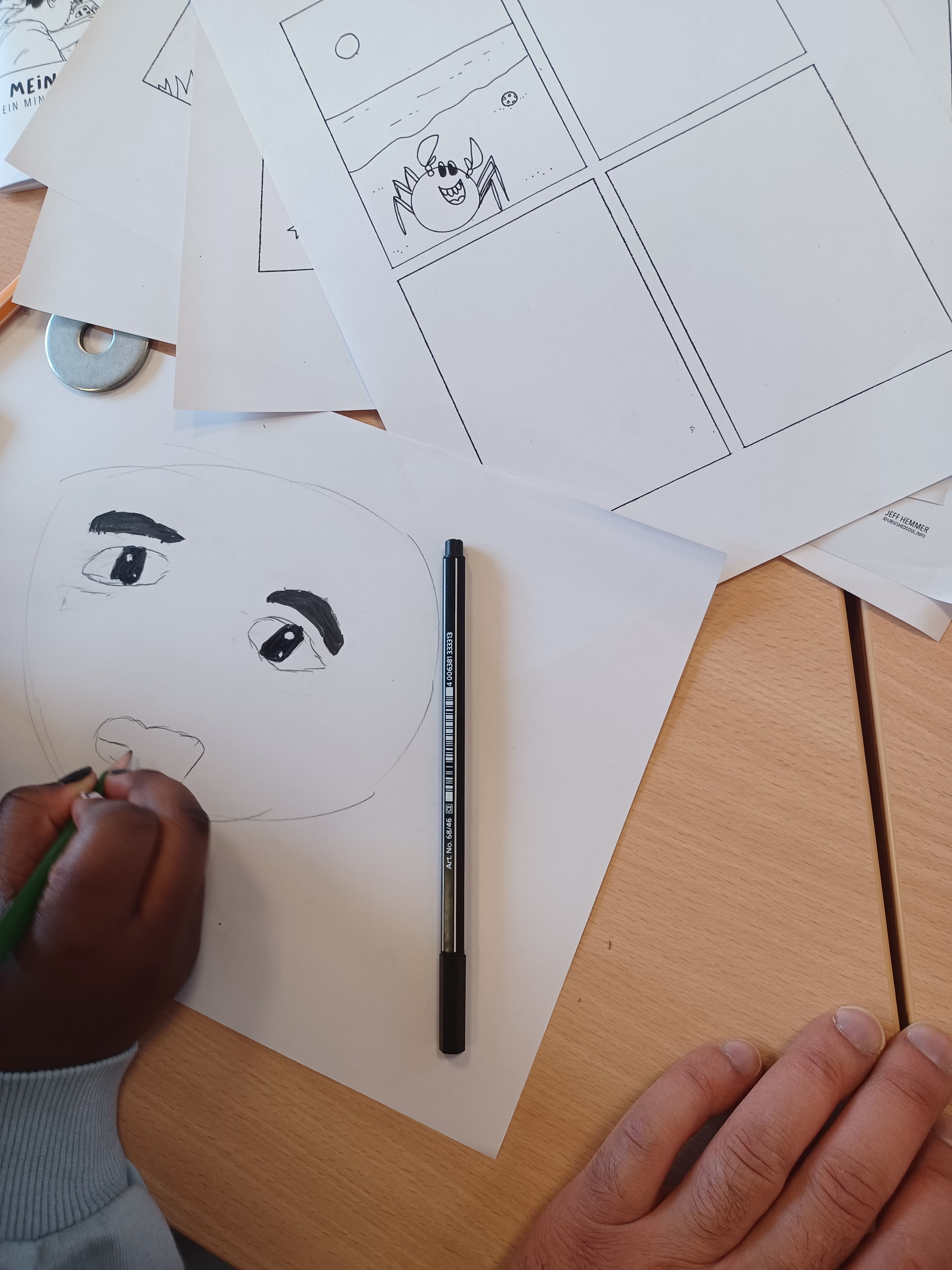 Auf einem Tisch liegen mehrere Blätter Papier. Man sieht die Hand eines schwarzen Kindes, das ein Gesicht zeichnet.